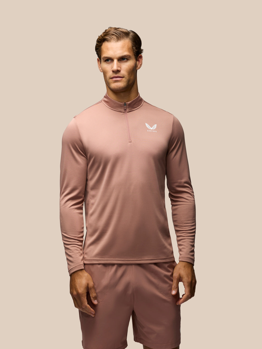 A man modeling a dusty rose long-sleeve athletic quarter-zip pullover with matching shorts against a solid beige background.