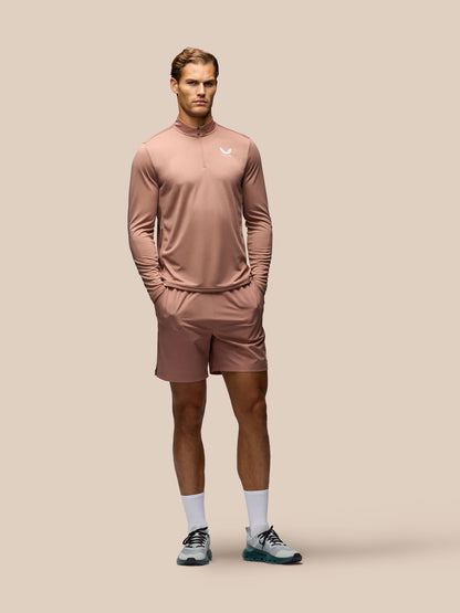 A male model stands against a solid beige studio background wearing a matching dusty rose athletic set consisting of a long-sleeve quarter-zip performance top and matching shorts, paired with white socks and gray running shoes.