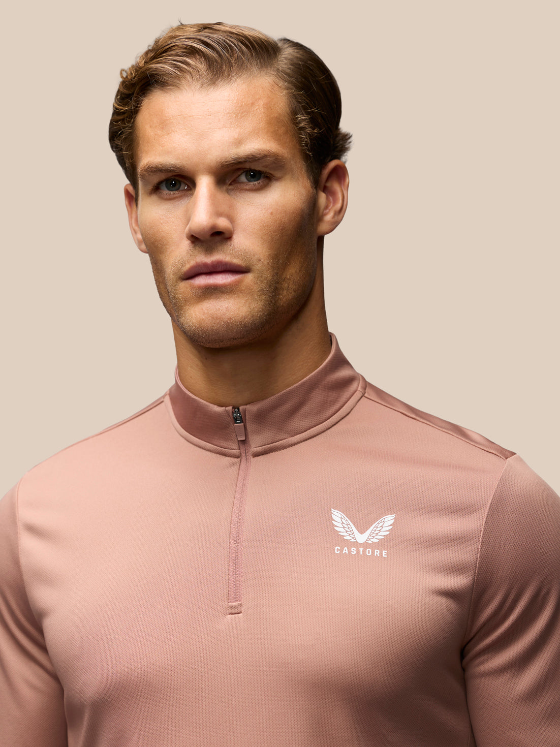 A man wearing a dusty rose quarter-zip athletic pullover with a white winged logo and the brand name 'CASTORE' on the left chest, set against a solid beige background.