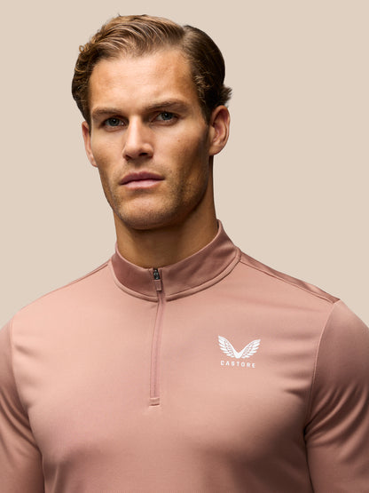 A man wearing a dusty rose quarter-zip athletic pullover with a white winged logo and the brand name 'CASTORE' on the left chest, set against a solid beige background.