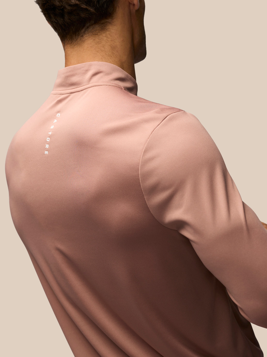 A close-up back view of a man wearing a dusty rose long-sleeve athletic quarter-zip shirt with a small vertical white logo on the upper back, set against a solid beige background.