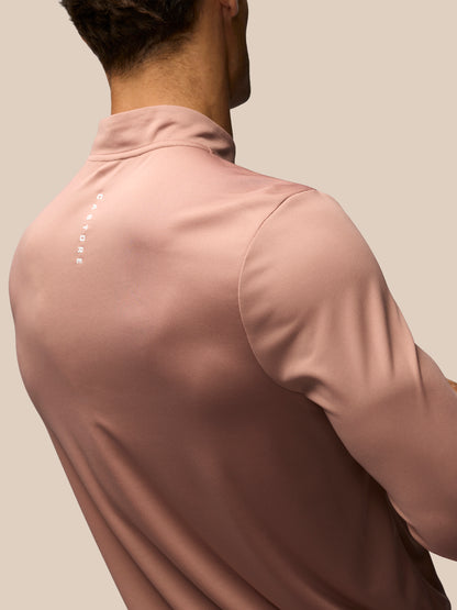 A close-up back view of a man wearing a dusty rose long-sleeve athletic quarter-zip shirt with a small vertical white logo on the upper back, set against a solid beige background.