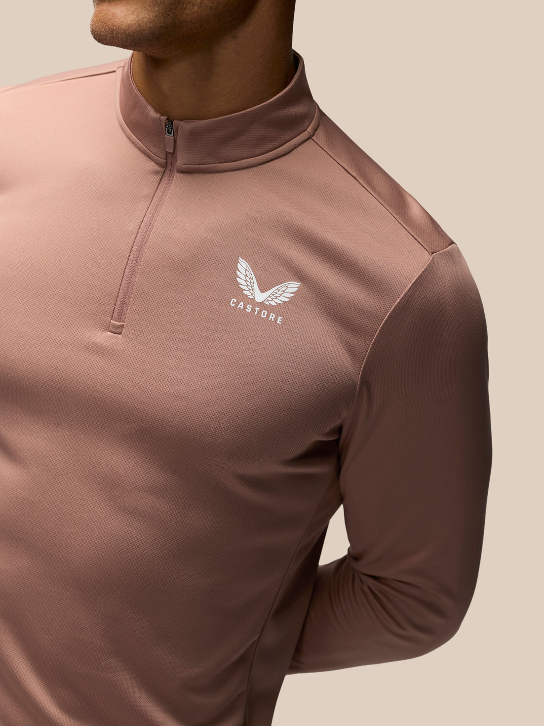 A close-up of a man wearing a dusty rose long-sleeve quarter-zip athletic top with a white winged logo and the brand name 'CASTORE' on the left chest, set against a solid beige background.
