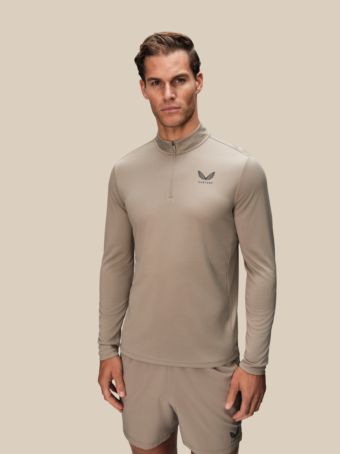 A man modeling a light taupe long-sleeve quarter-zip athletic top with a small dark logo on the left chest, paired with matching shorts, against a solid light beige background.