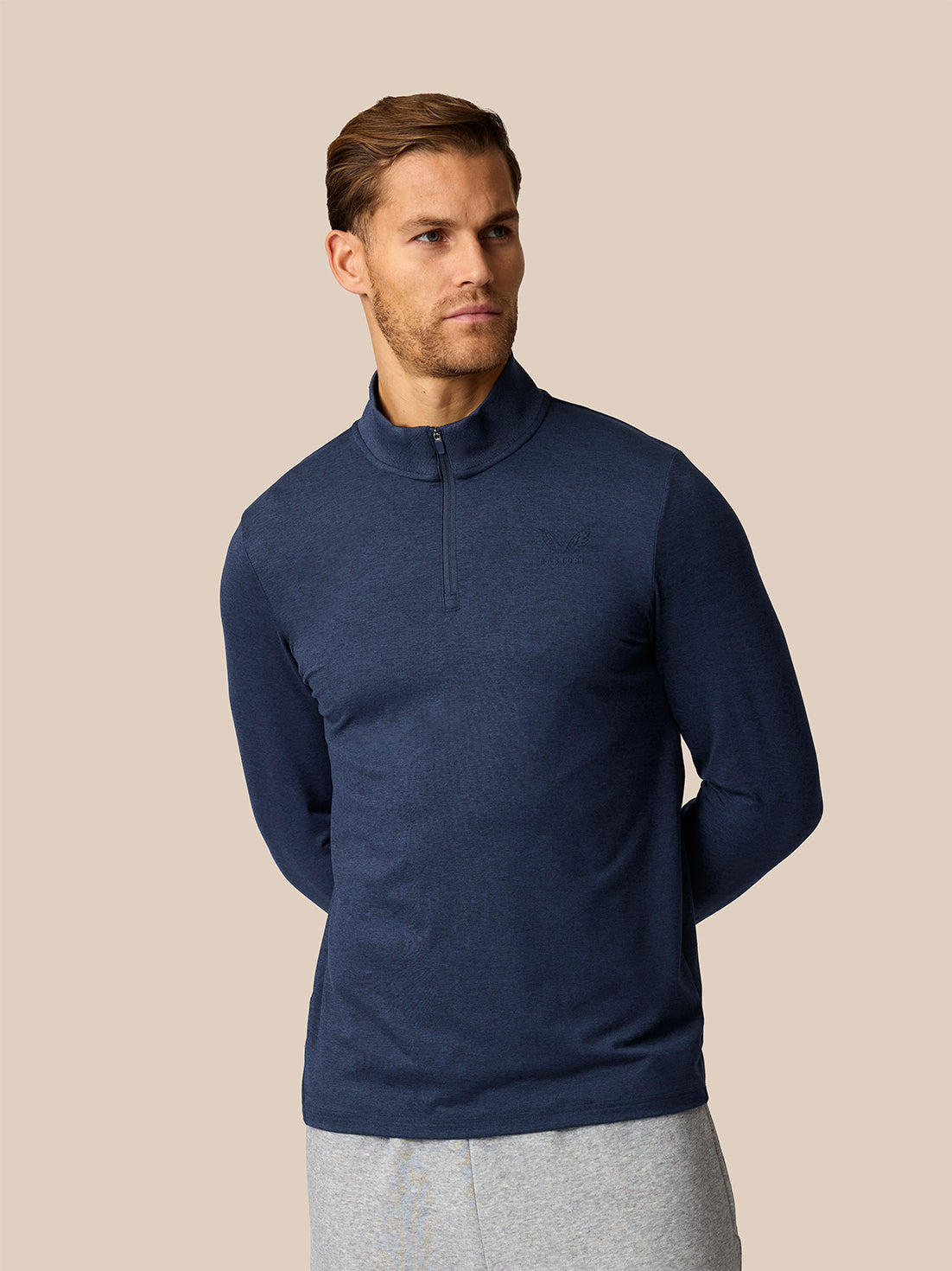 A man wearing a navy blue quarter-zip mock-neck long-sleeve shirt and light gray sweatpants, standing against a solid beige background.