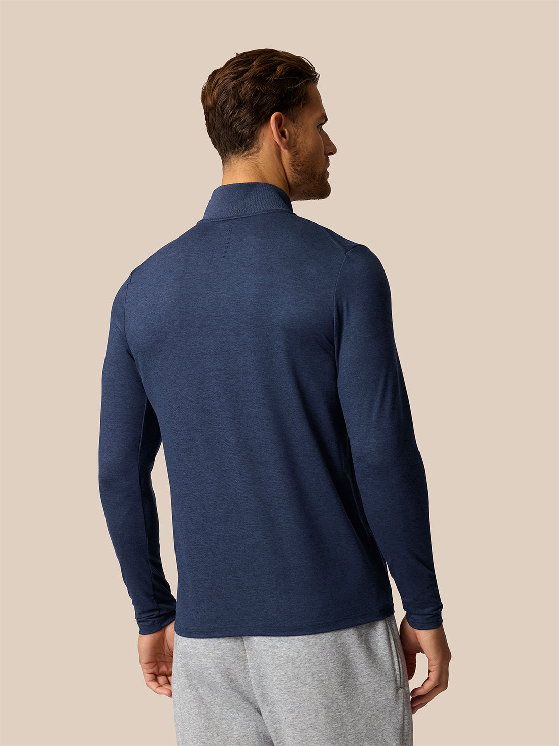 A man shown from the back wearing a navy blue long-sleeve mock neck shirt and light gray sweatpants, set against a solid beige background.
