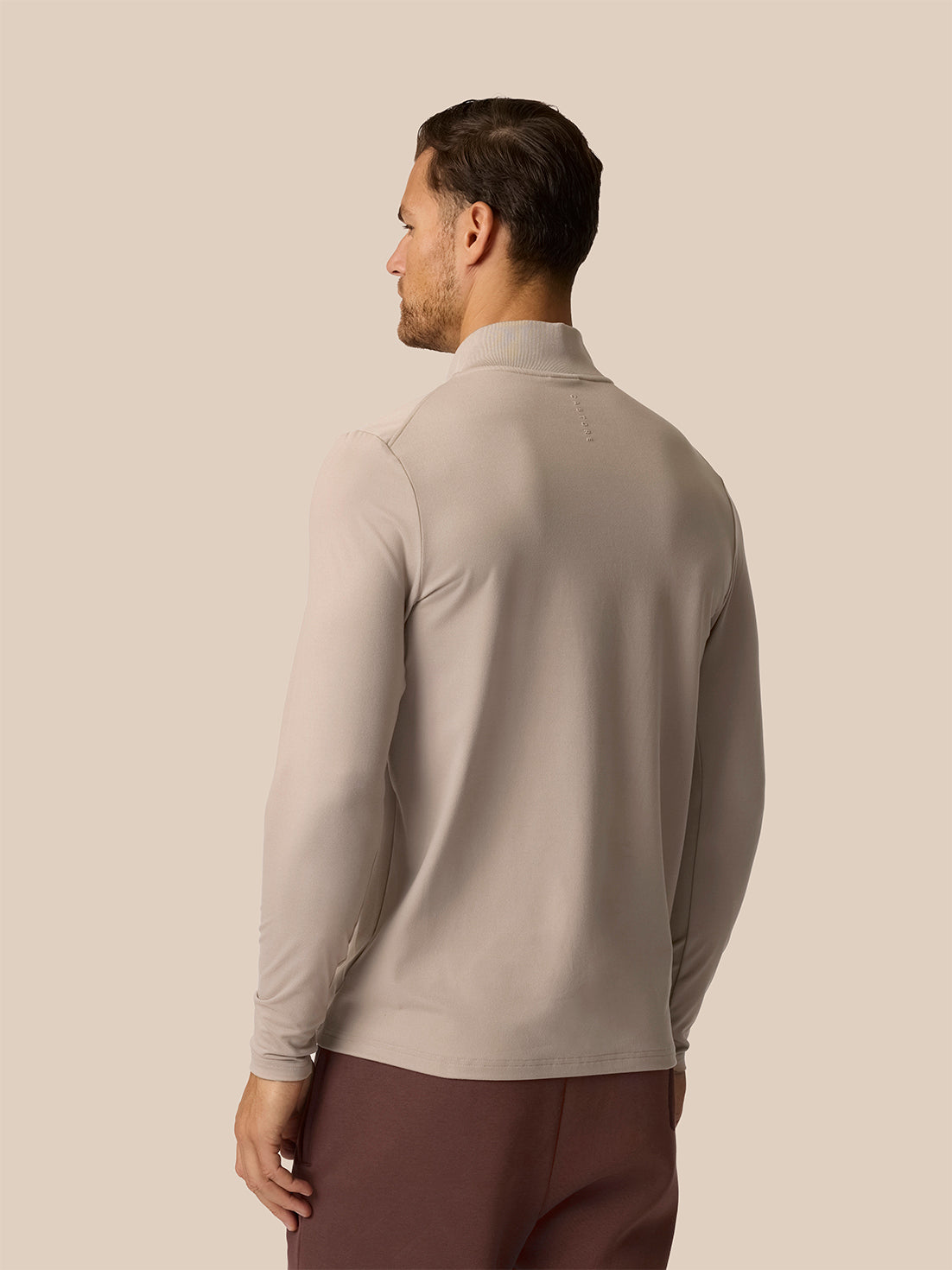 A man shown from the back wearing a light taupe long-sleeve mock neck shirt and dark brown trousers, set against a solid beige background.