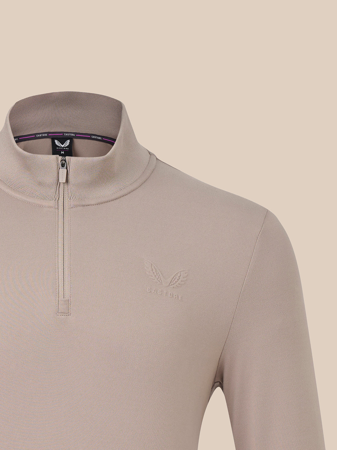 Close-up of a beige quarter-zip pullover top with a mock neck collar and a subtle winged logo on the left chest, shown against a matching beige background.