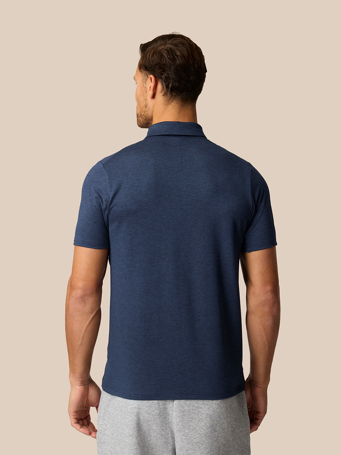 A man viewed from the back wearing a navy blue short-sleeve polo shirt and light gray sweatpants, set against a solid beige background.