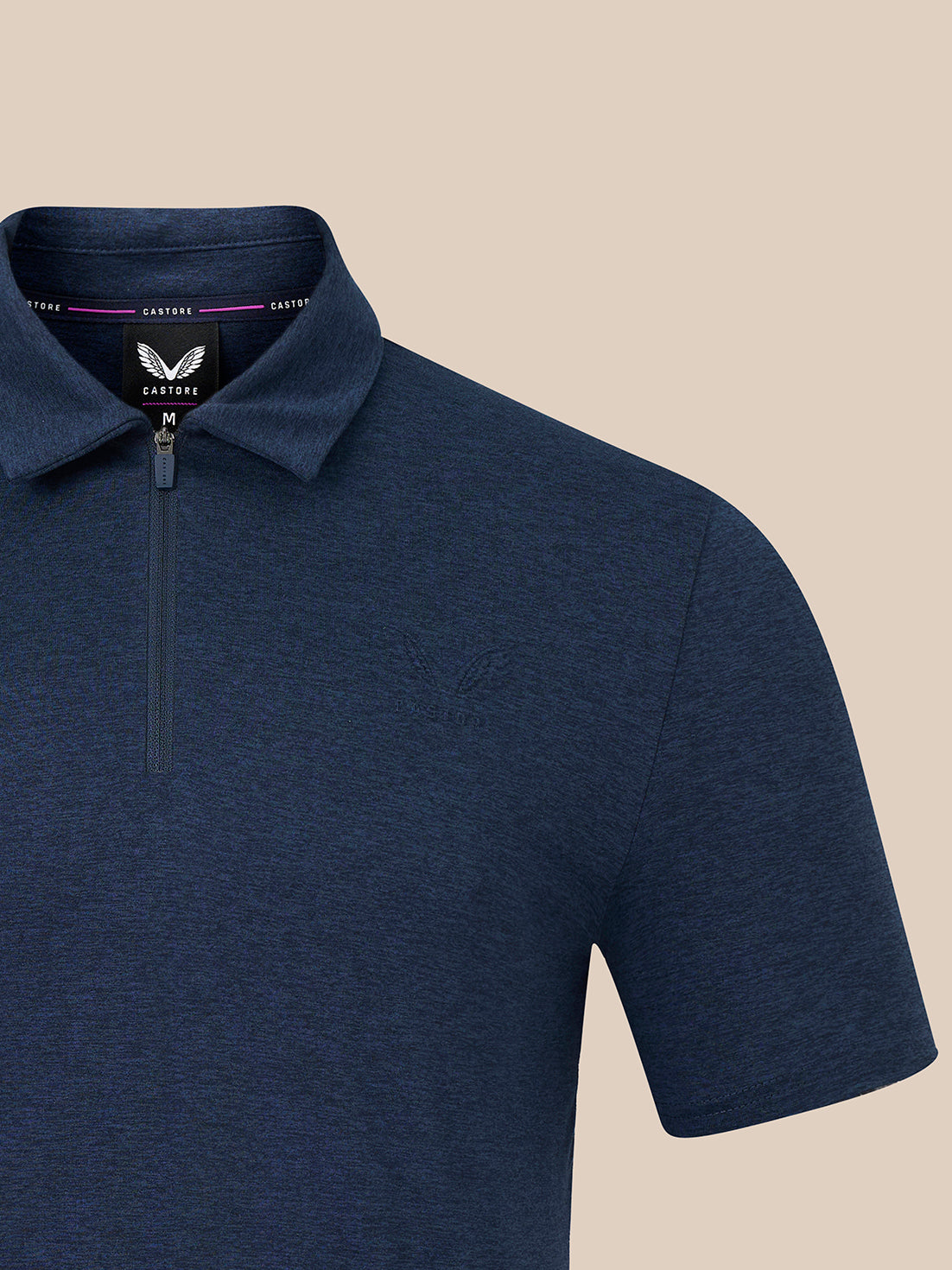 Close-up of a navy blue short-sleeve polo shirt with a half-zip collar, shown against a solid beige background. The brand label inside the collar reads Castore.