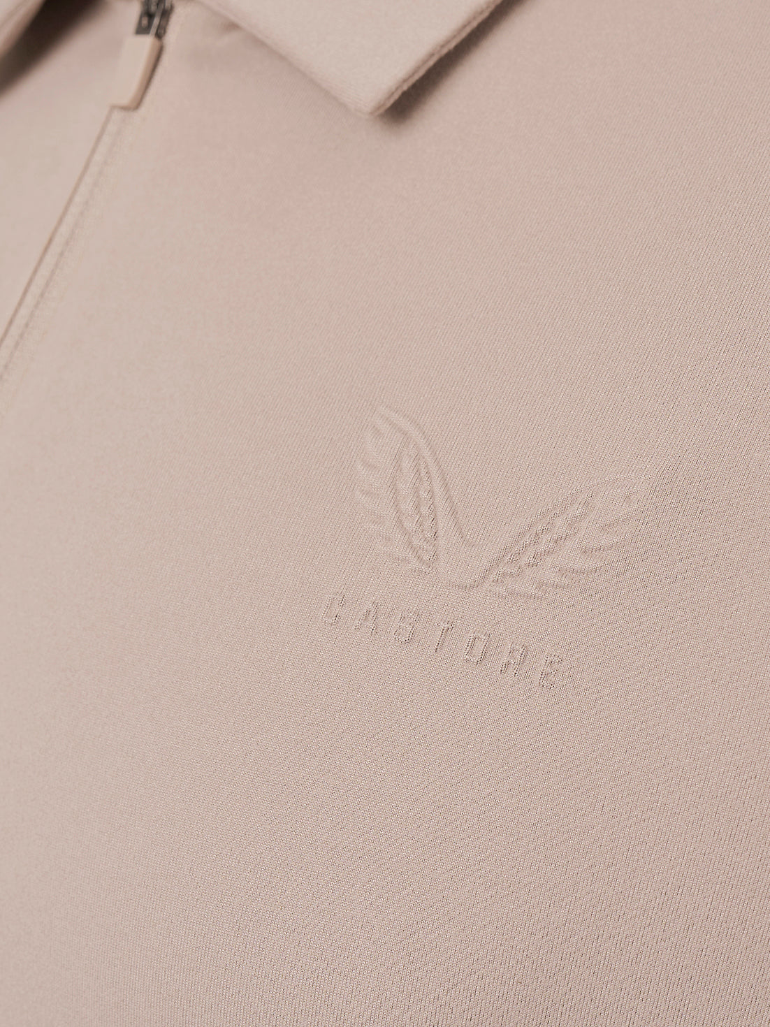 Close-up of a light beige zip-up jacket or sweater showing the collar, zipper pull, and an embossed winged logo with the word CASTORE on the chest.