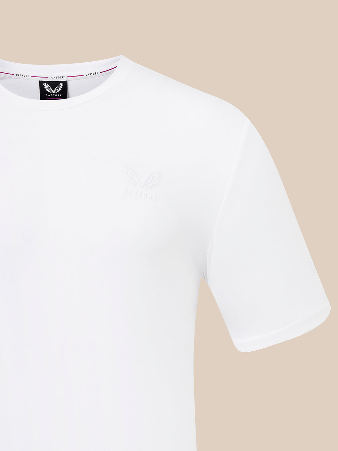 A close-up of a white short-sleeve crewneck t-shirt by CASTORE, featuring a subtle tonal embroidered logo on the left chest and a black brand label inside the collar, set against a solid beige background.