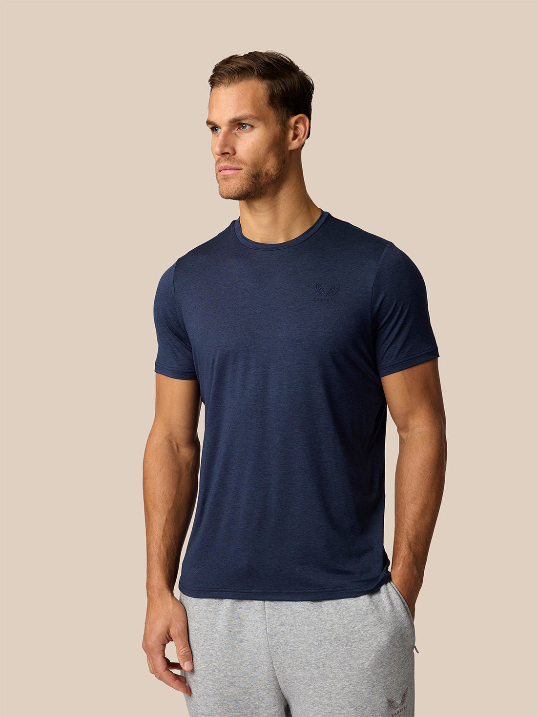 A man wearing a navy blue short-sleeve crewneck t-shirt and light gray sweatpants, standing against a solid beige background.