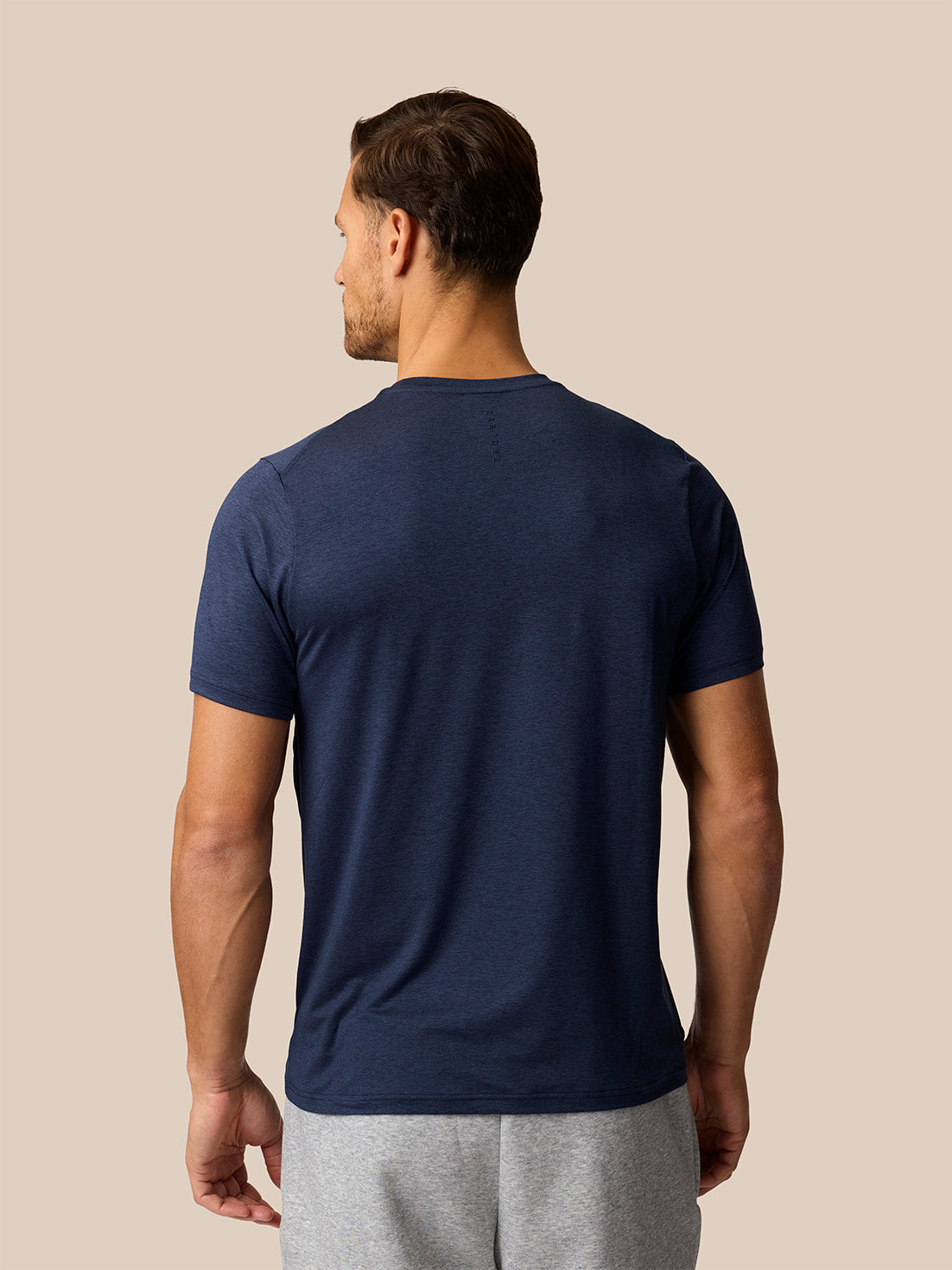 A man viewed from the back wearing a navy blue short-sleeve crewneck t-shirt and light gray sweatpants, set against a solid beige background.