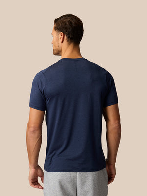 A man viewed from the back wearing a navy blue short-sleeve crewneck t-shirt and light gray sweatpants, set against a solid beige background.