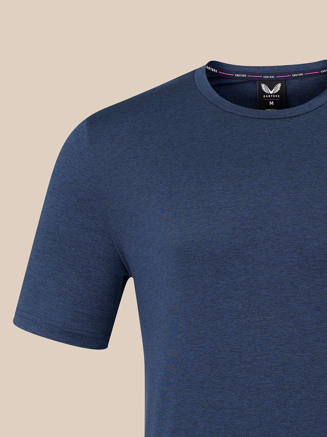 A close-up of a short-sleeve crew-neck t-shirt in heathered navy blue, shown on a light beige background. The inner neck label features a logo with a winged logo and the brand name 'Castore'.