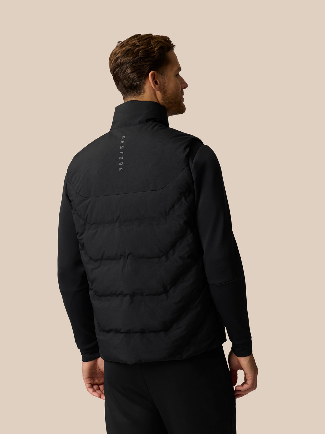 A man shown from the back wearing a black puffer vest over a black long-sleeve top, against a solid beige background. The brand name 'CASTORE' is printed vertically in white on the upper back of the vest.