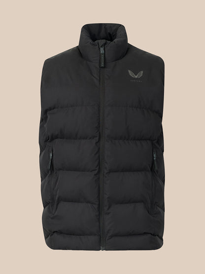 A black puffer vest with a stand-up collar and a small dark gray wing logo on the left chest, displayed against a solid beige background.