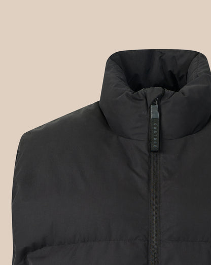 Close-up of a black puffer vest with a high stand-up collar and a black zipper pull tab featuring the brand name 'CASTORE' in white text, set against a solid beige background.