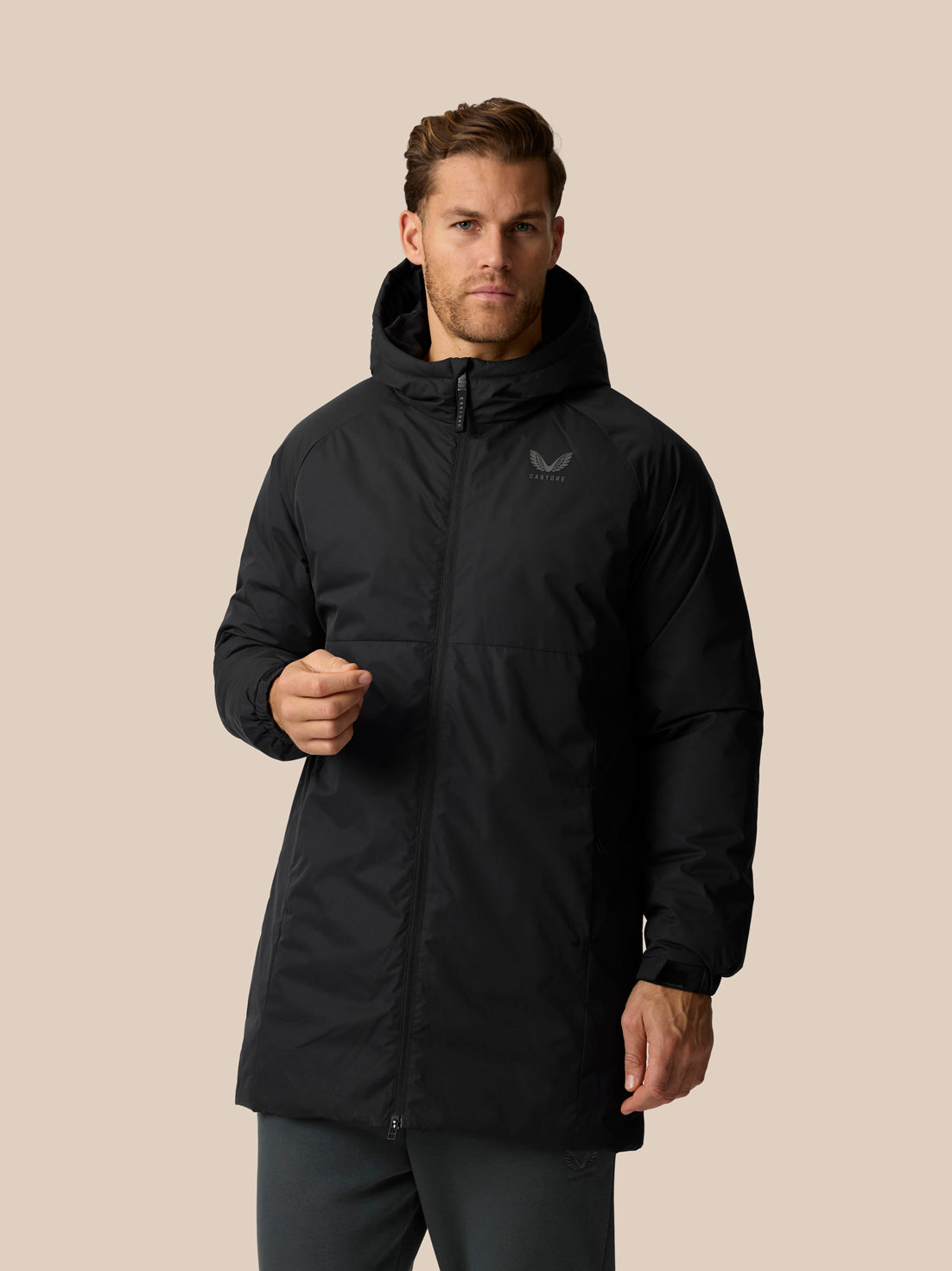 A man wearing a black hooded puffer jacket with a subtle wing logo on the left chest, posed against a solid beige background.