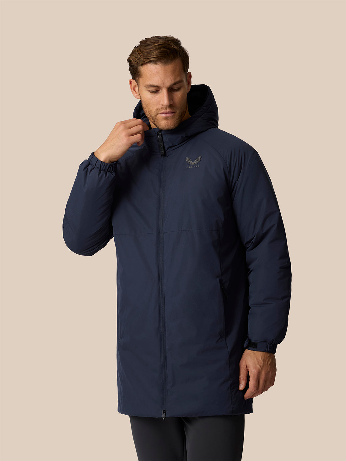 A man wearing a navy blue hooded puffer jacket with a small embroidered logo on the left chest, posed against a solid beige background.