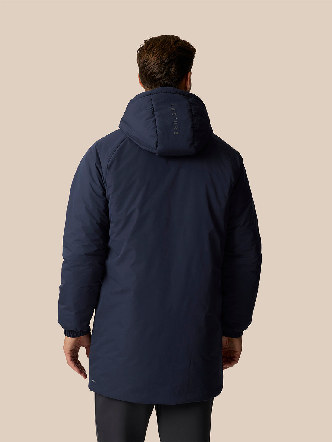 A man viewed from the back wearing a navy blue hooded puffer jacket against a solid beige background.