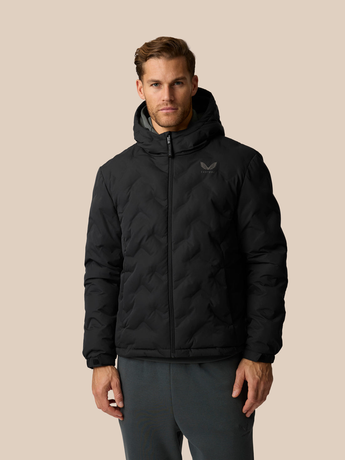 A man wearing a black hooded puffer jacket with a subtle tonal logo on the left chest, paired with dark gray pants, against a solid beige background.