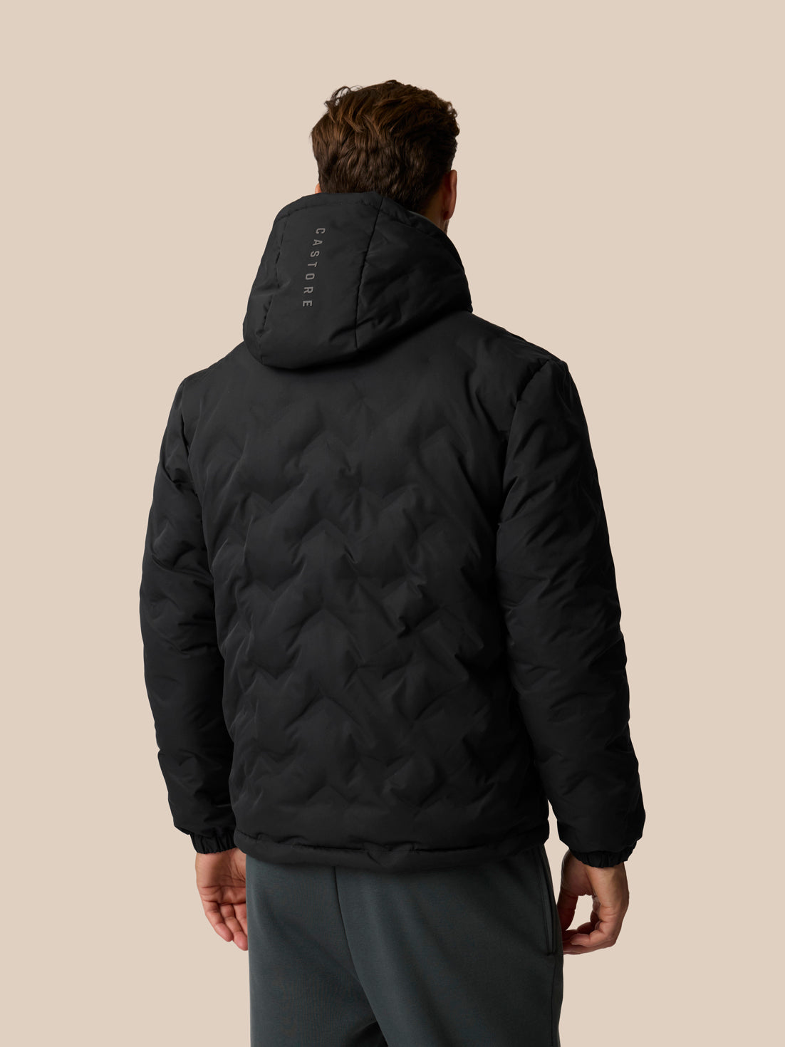 A man viewed from behind wearing a black quilted puffer jacket with a hood and a visible vertical 'CASTORE' logo on the back, paired with dark gray pants, against a solid beige background.