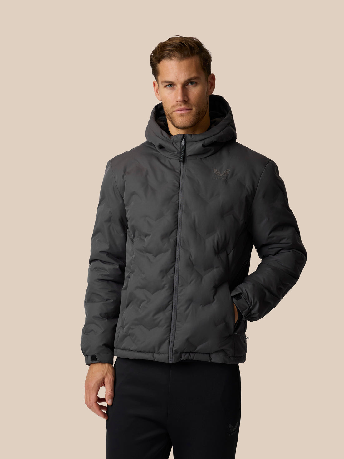 A man wearing a dark gray hooded puffer jacket with a full-zip front, photographed against a solid beige background.