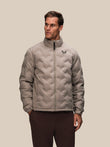 Man wearing a taupe padded quilted jacket with a stand up collar and Castore logo on the chest.
