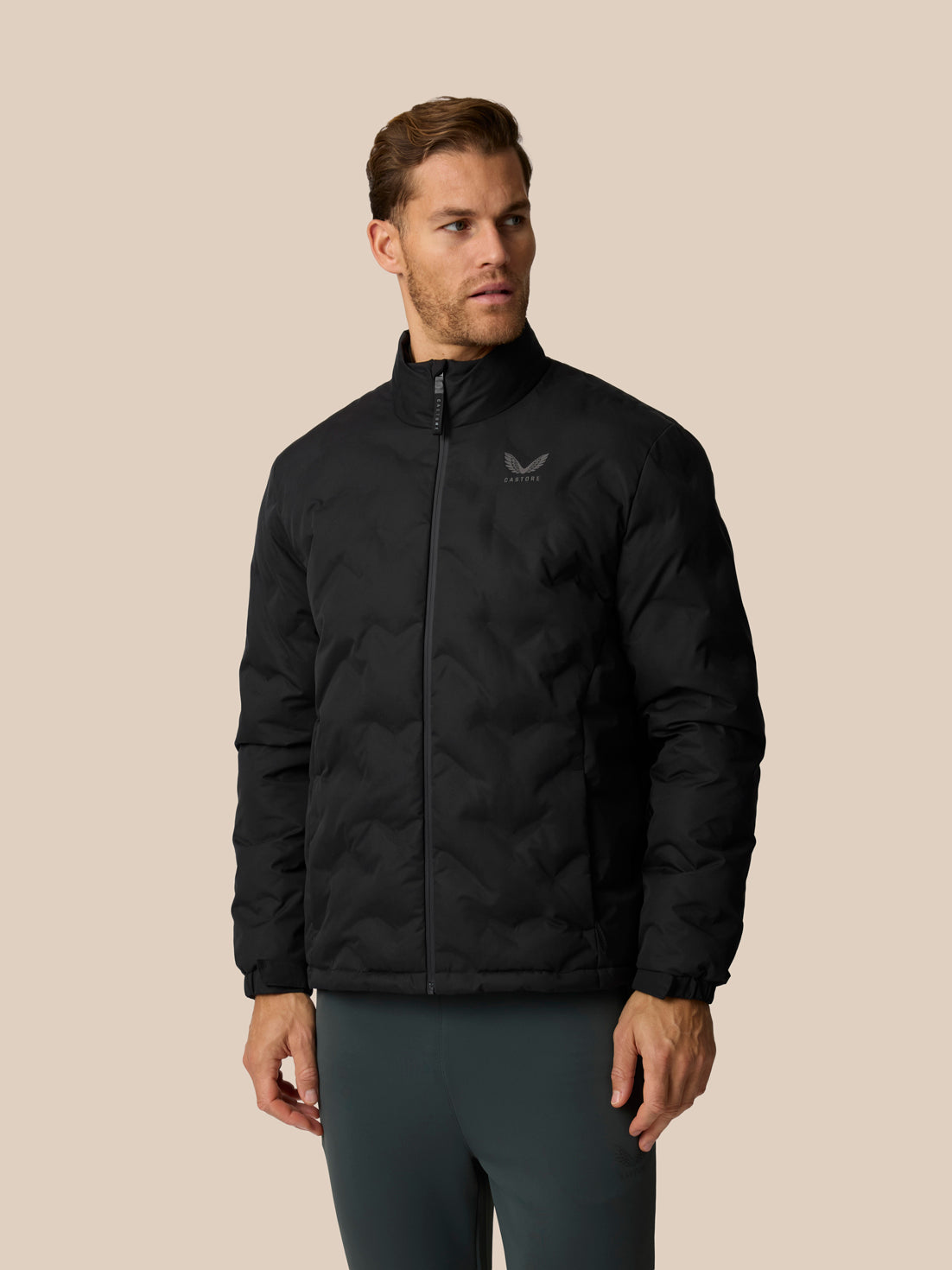 A man wearing a black quilted zip-up jacket with a small gray wing logo on the left chest, paired with dark gray leggings, set against a solid beige background.