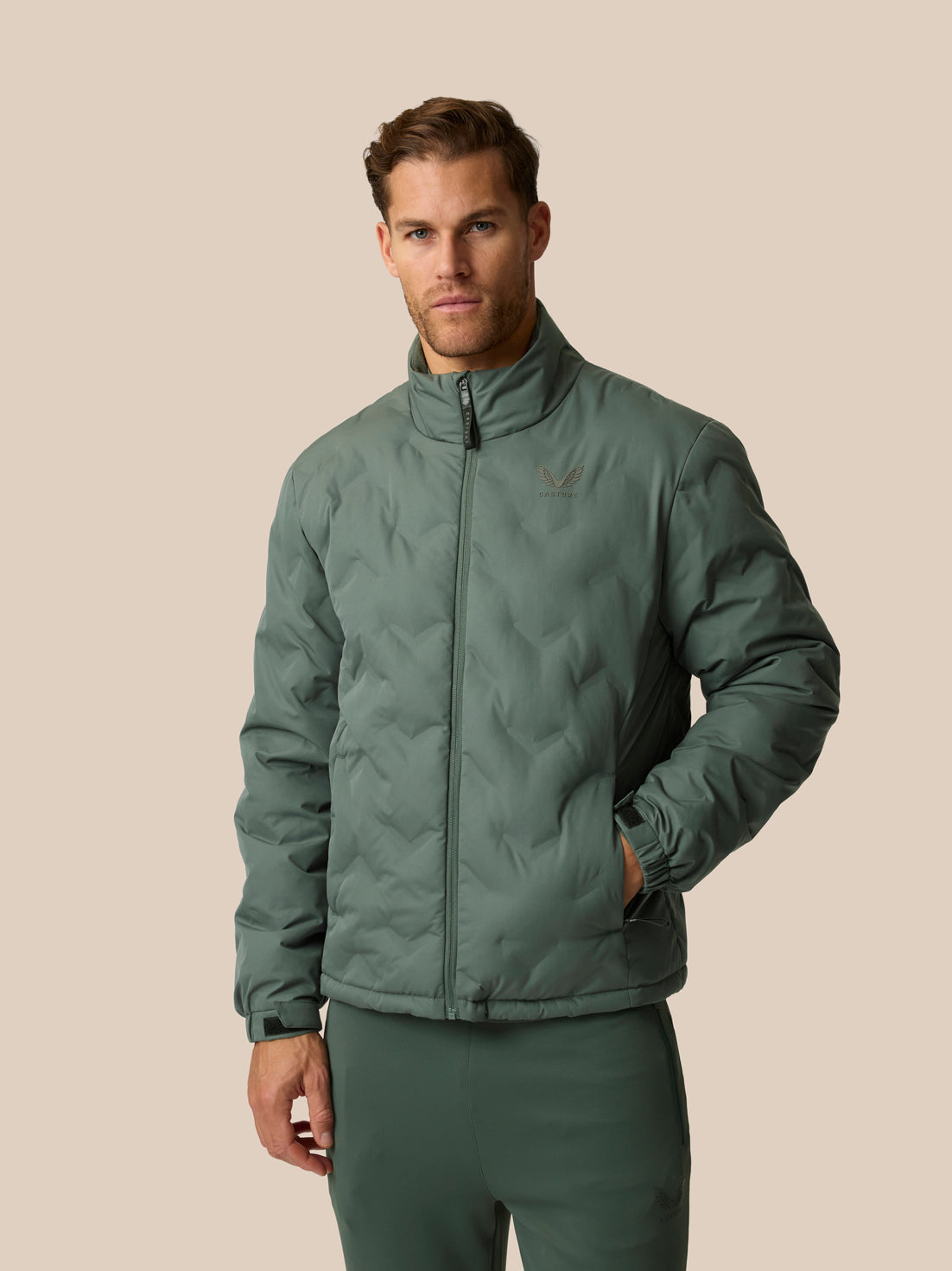 A man wearing a sage green quilted zip-up jacket and matching green pants, standing against a solid beige background.
