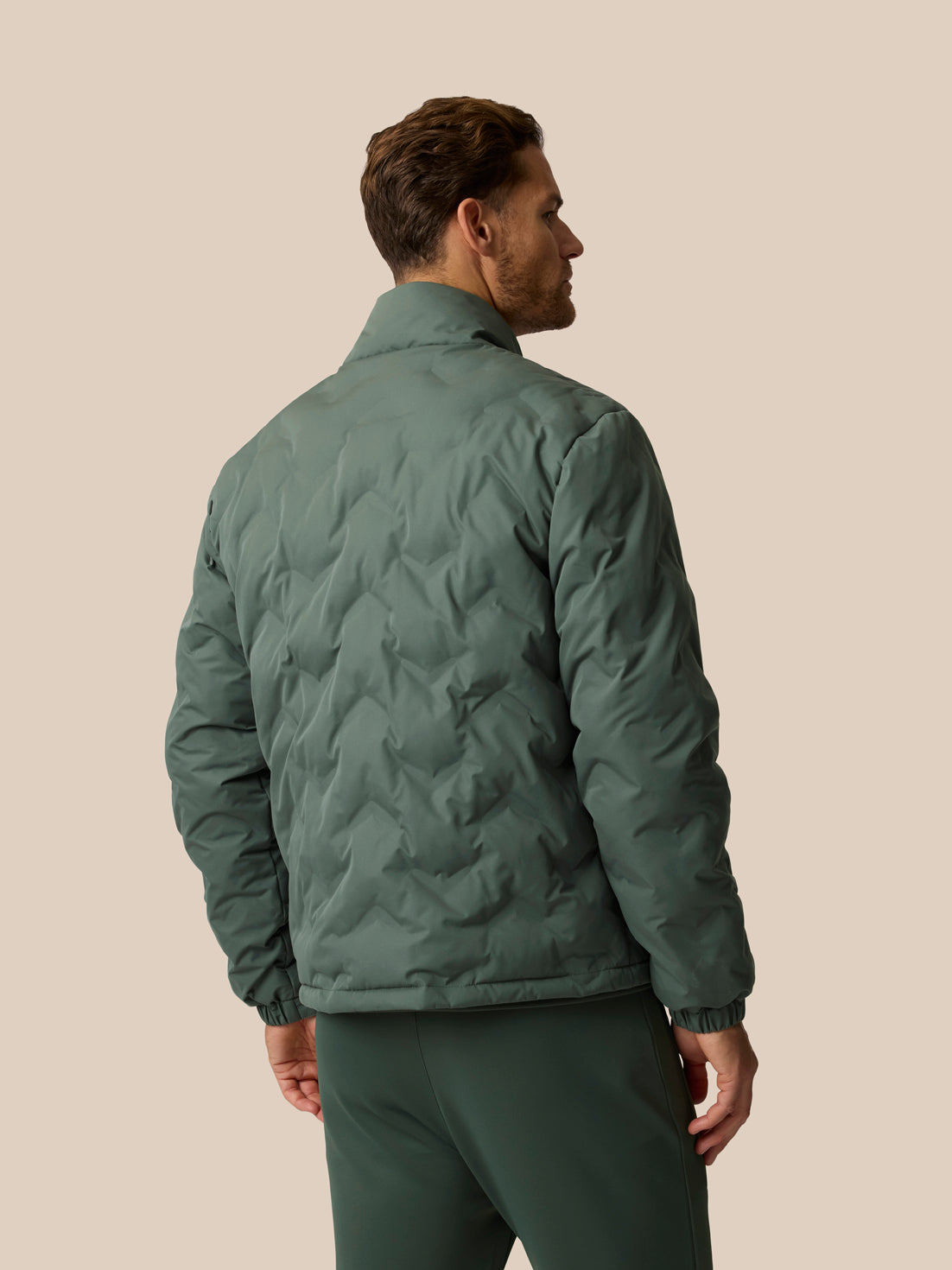 A man shown from the back wearing a dark green quilted jacket and matching green pants, standing against a solid beige background.