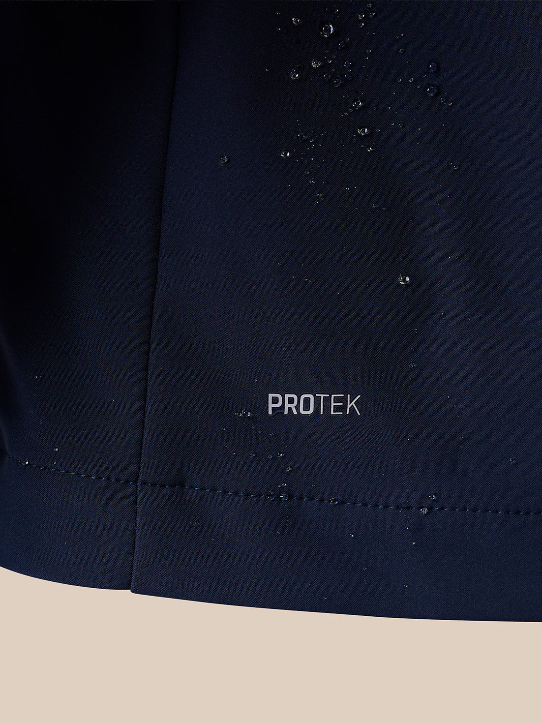 Close-up of a dark navy fabric surface with water droplets beaded on it and the word PROTEK printed near the center, set against a light beige background.