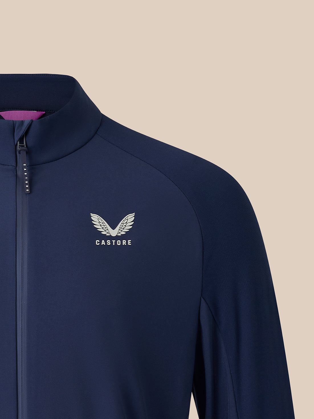 Close-up of a navy blue zip-up athletic jacket with a white Castore wing logo on the chest, set against a solid beige background.