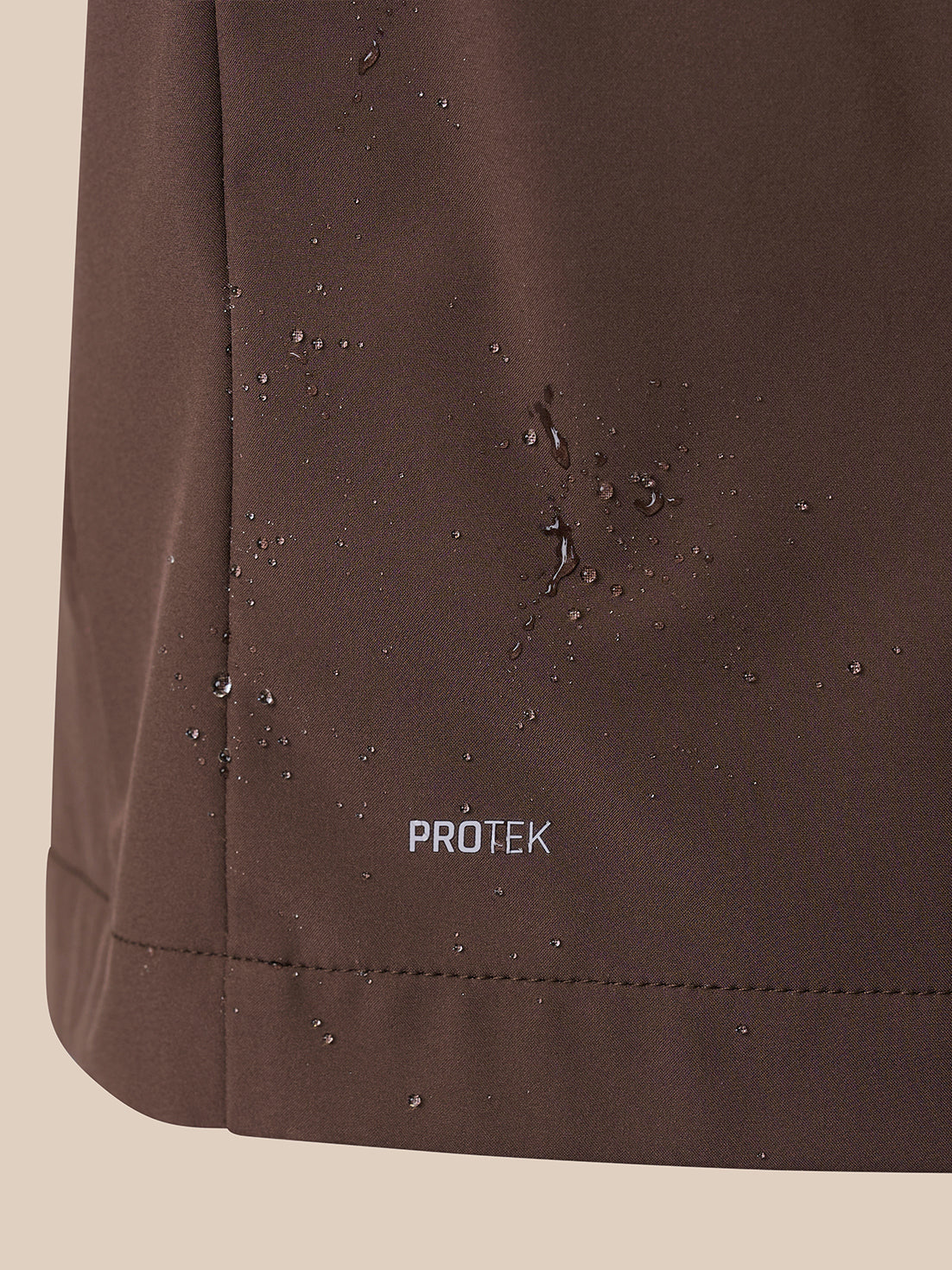 Close-up of a dark brown waterproof fabric surface with water droplets beading on it and the word PROTEK printed near the bottom.