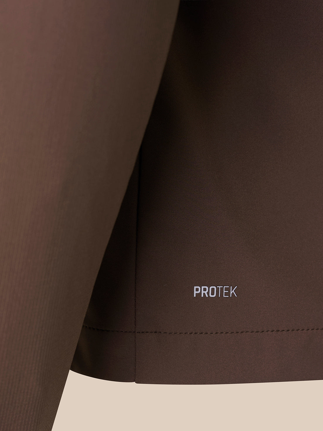 Close-up of a dark brown athletic garment showing the side seam and hem with the word PROTEK printed in small white text.