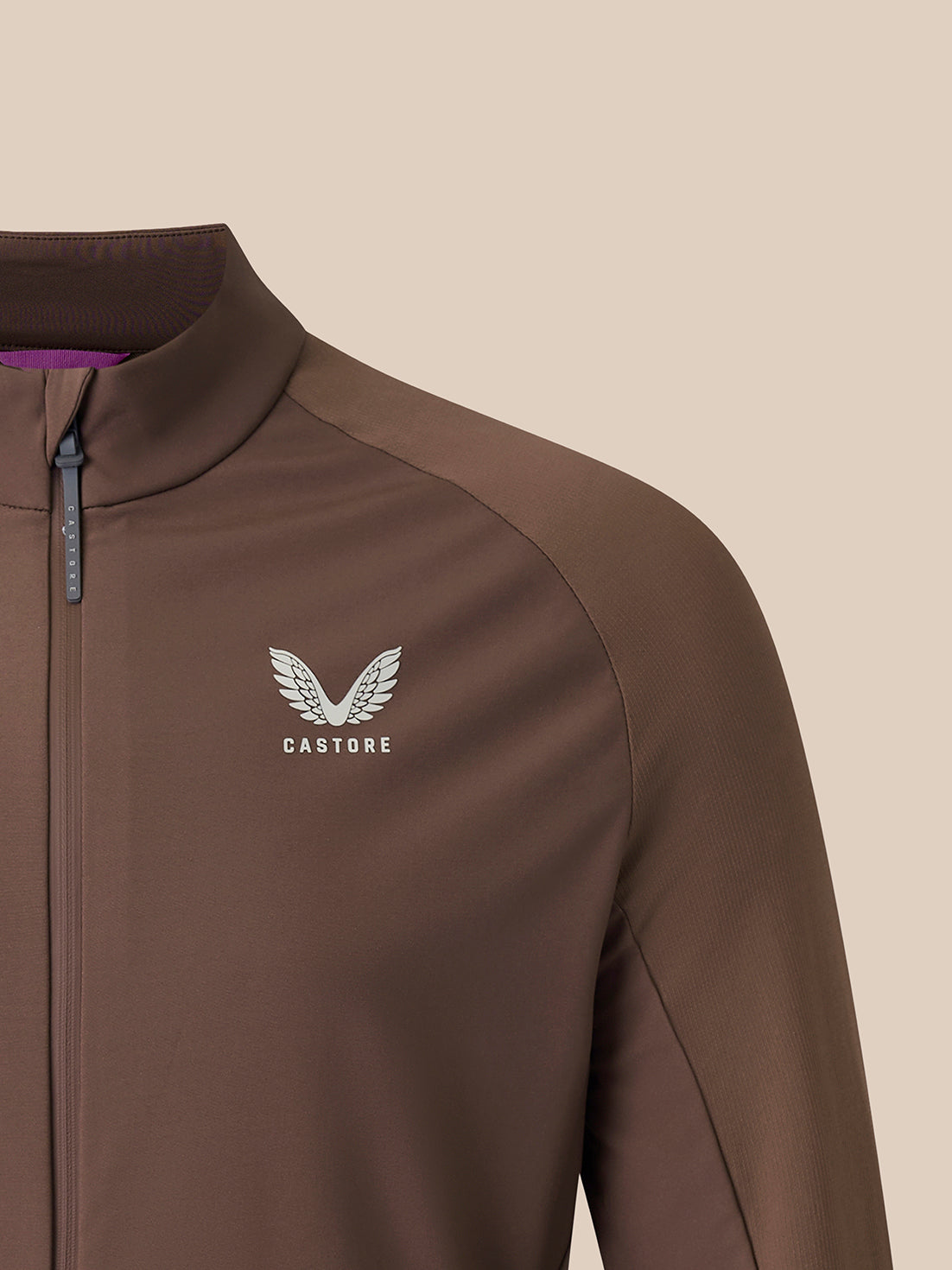 A close-up of a brown athletic zip-up jacket with a stand collar, featuring a silver winged Castore logo on the left chest and a purple inner collar lining.