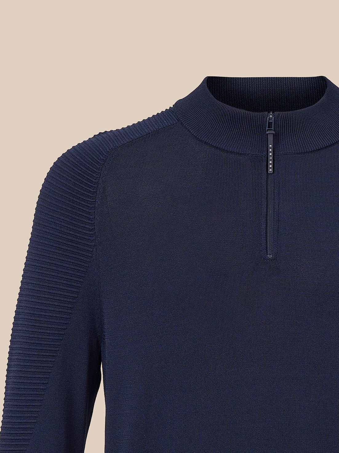 Close-up of a navy blue ribbed knit sweater with a quarter-zip mock neck and textured shoulder panels, shown against a solid beige background.