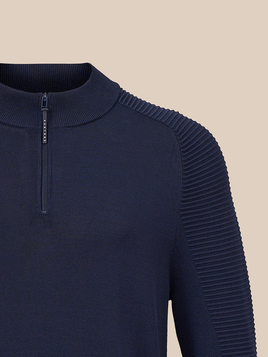Close-up of a navy blue ribbed knit quarter-zip sweater with pleated shoulder detailing, shown against a solid beige background.