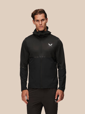 A man wearing a black hooded athletic jacket with a white wing logo on the left chest, standing against a solid beige background.