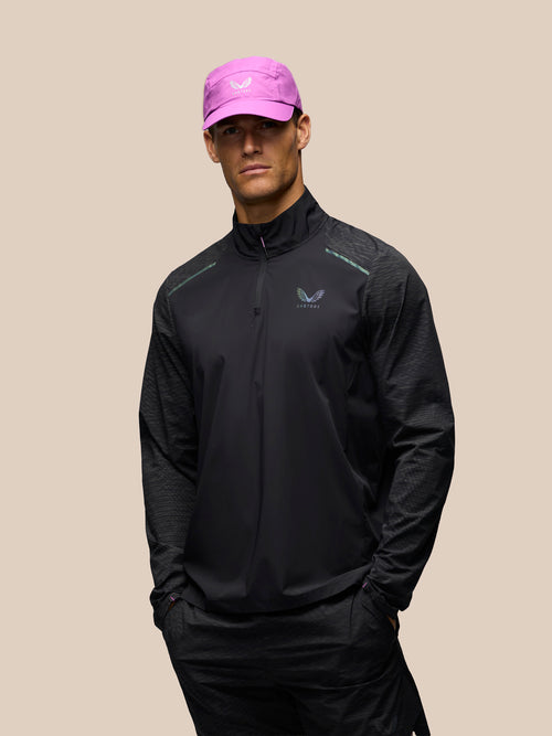A man wearing a black athletic zip-up jacket and matching black pants, paired with a bright pink cap, standing against a solid beige background.