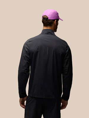 A man viewed from behind wearing a black long-sleeve athletic jacket and a bright pink cap, set against a solid beige background.
