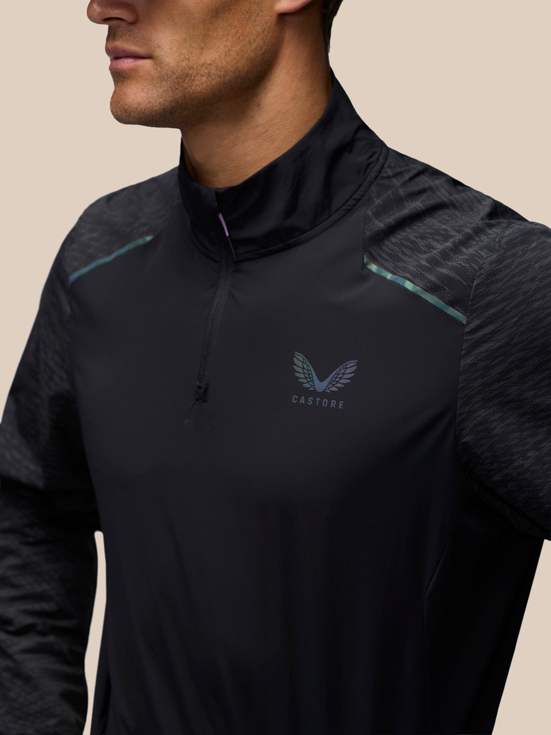 A close-up of a man wearing a black athletic quarter-zip pullover with a subtle wing logo and the brand name CASTORE on the chest, set against a solid beige background.