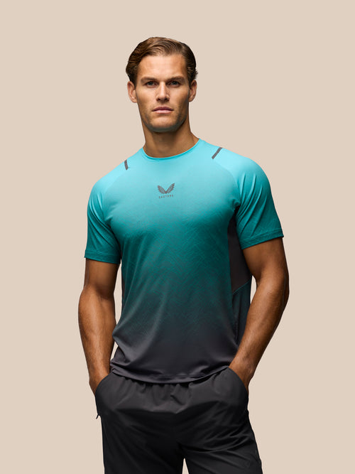 A man wearing a teal and dark gray gradient athletic t-shirt with a small wing logo on the chest, paired with dark gray athletic shorts, set against a solid beige background.
