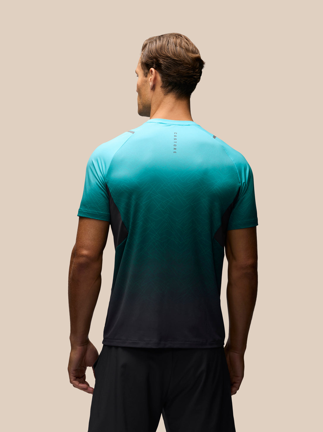 A man viewed from the back wearing a short-sleeve athletic shirt with a teal-to-black gradient, paired with black shorts, against a solid beige background.