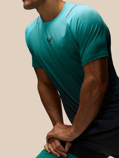 A close-up of a man wearing a teal athletic t-shirt with a subtle gradient fading to black at the sides, paired with dark athletic shorts, set against a solid beige background.