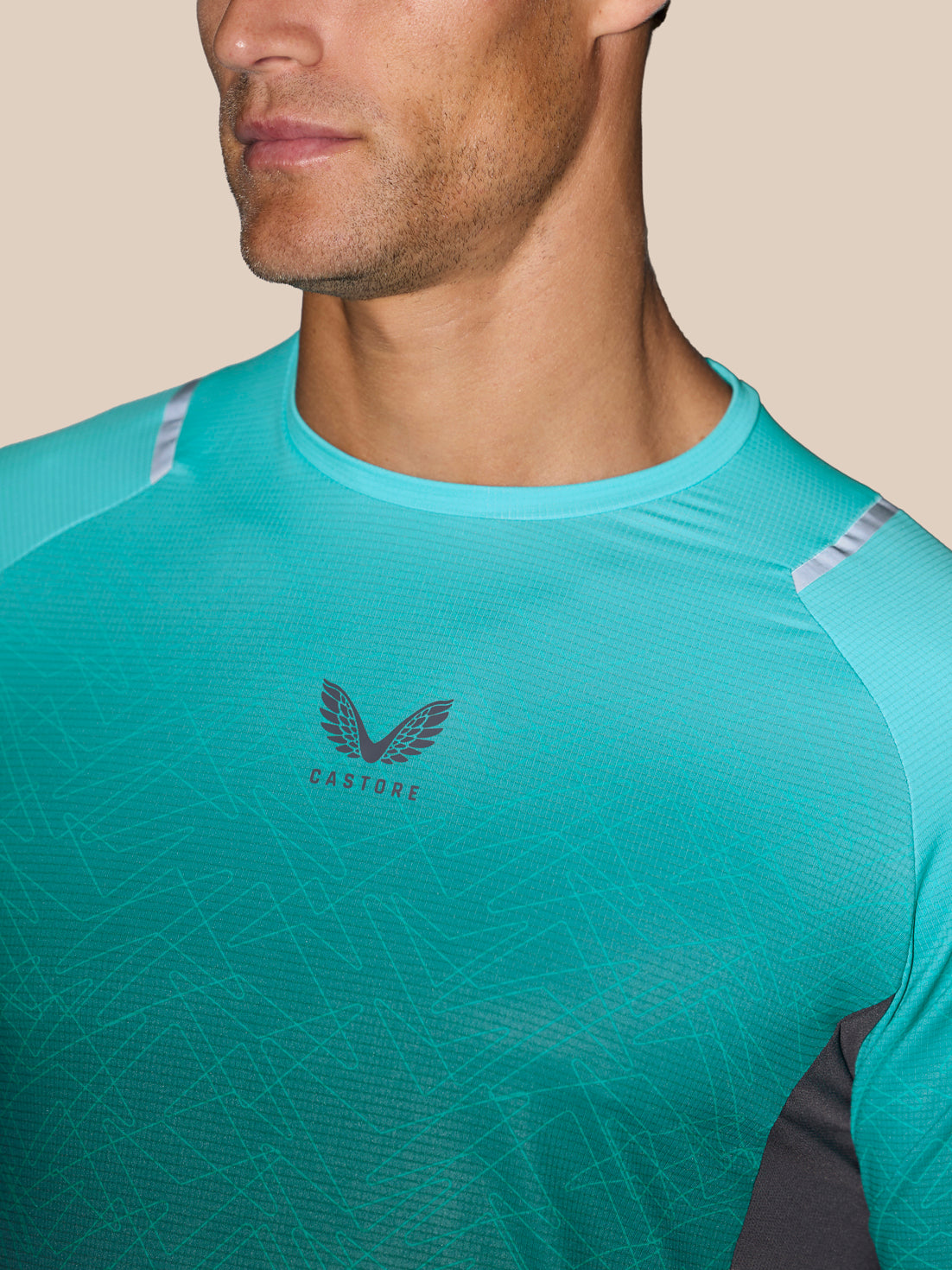 A close-up of a man wearing a turquoise athletic t-shirt with a dark gray winged logo and the word 'CASTORE' on the chest, set against a solid beige background.