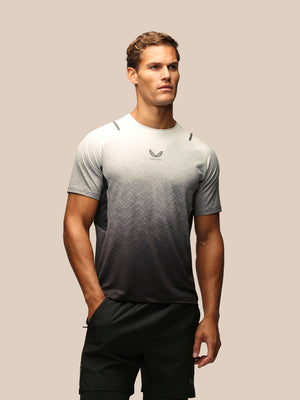 A man modeling a short-sleeve athletic t-shirt with a gray-to-black gradient and subtle geometric pattern, paired with black shorts, against a solid beige background.