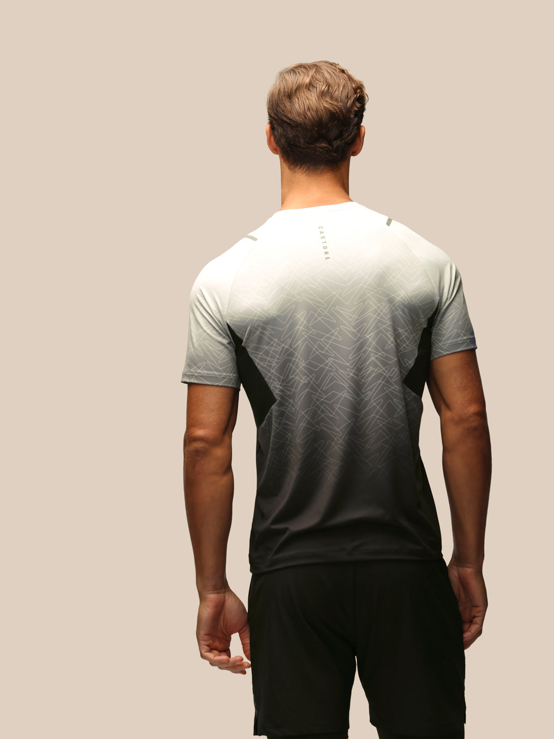 A man viewed from behind wearing a short-sleeve athletic t-shirt with a gradient design that fades from white at the shoulders to black at the waist, paired with black shorts against a solid beige background.